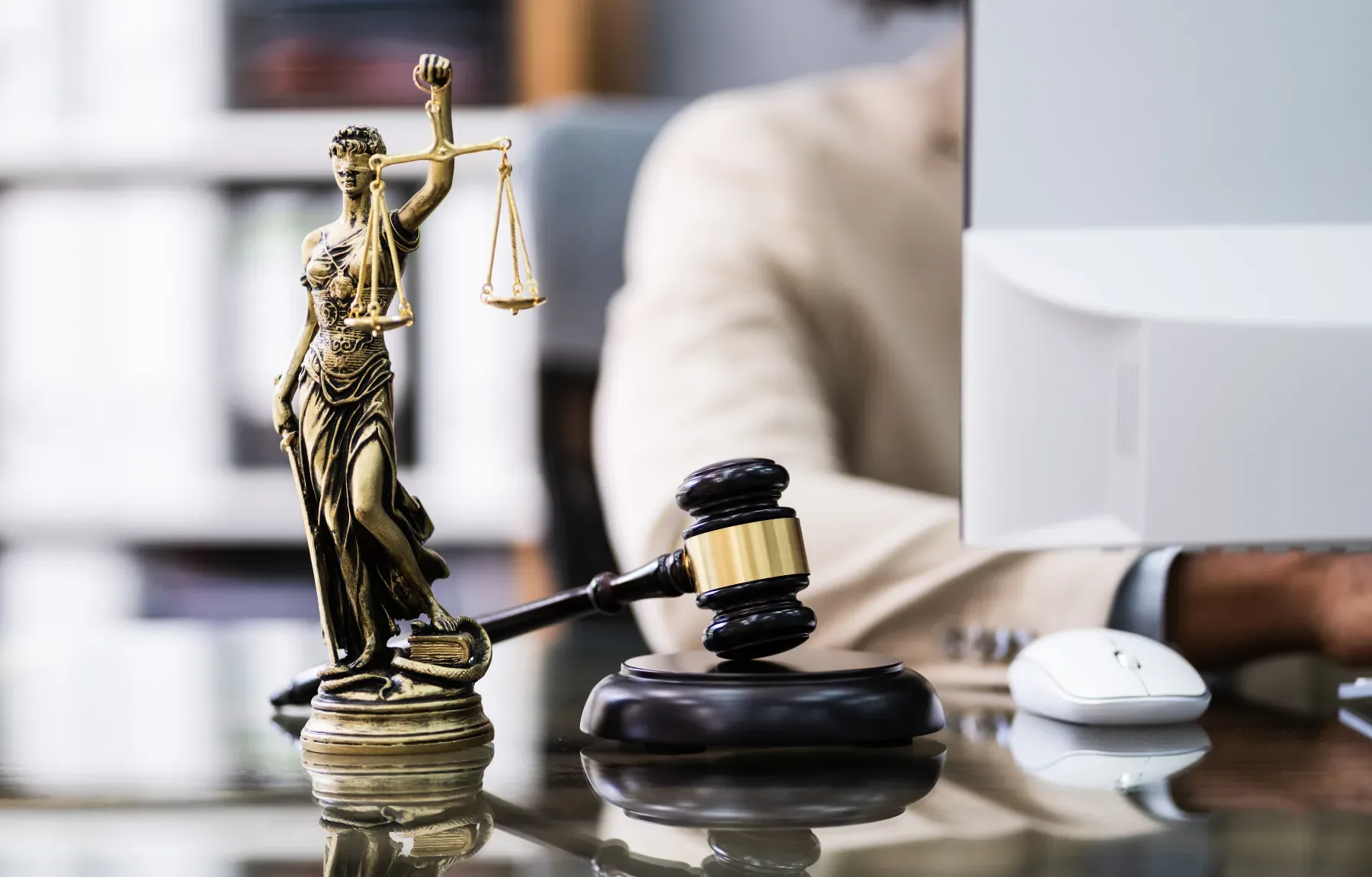 A brass Lady Justice statuette, one arm raised holding a set of balanced scales, stands in sharp focus beside a dark wood gav